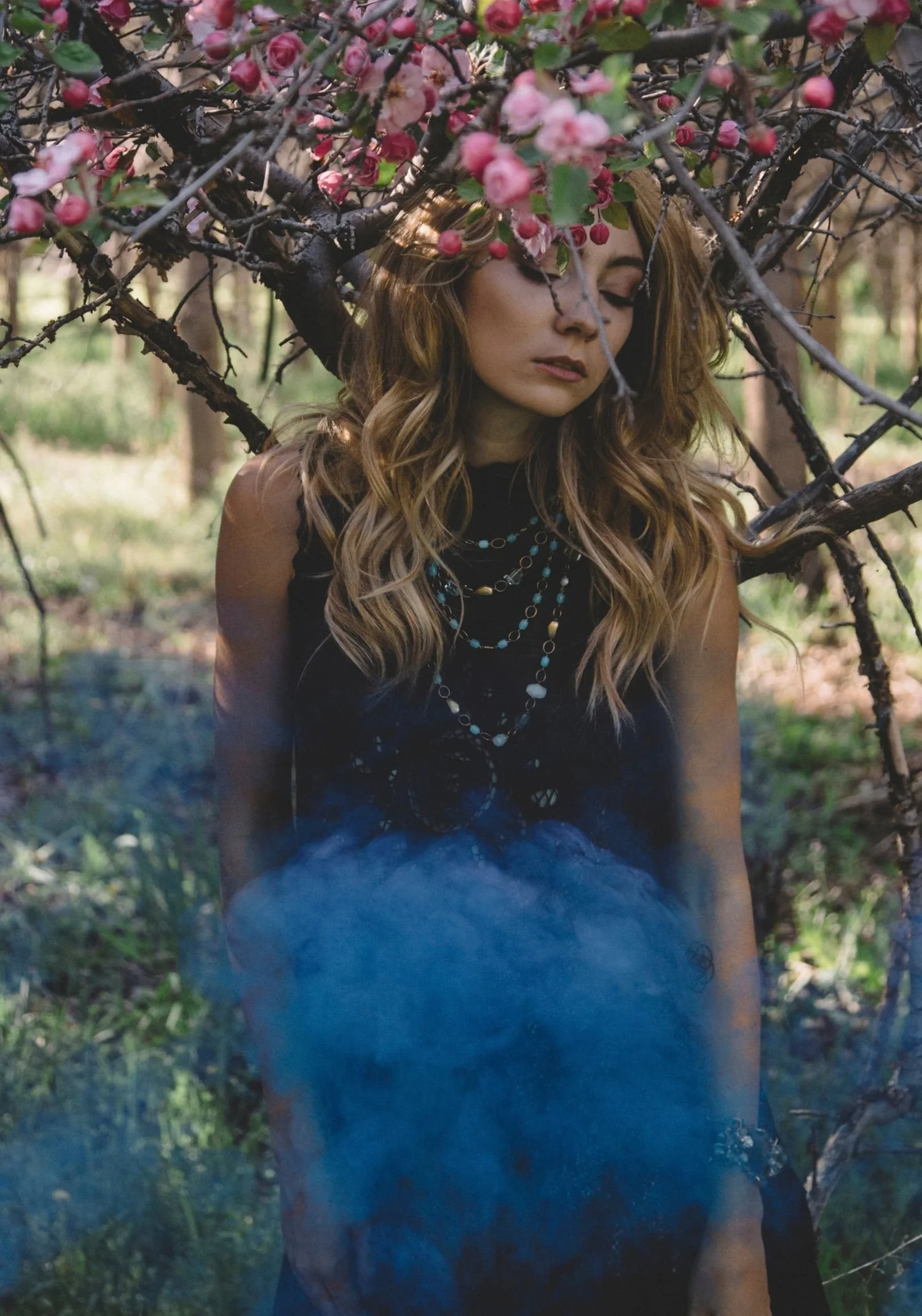 A model in a black dress under a canopy of pink blossoms, blue smoke rising at her waist, wearing layered gemstone necklaces