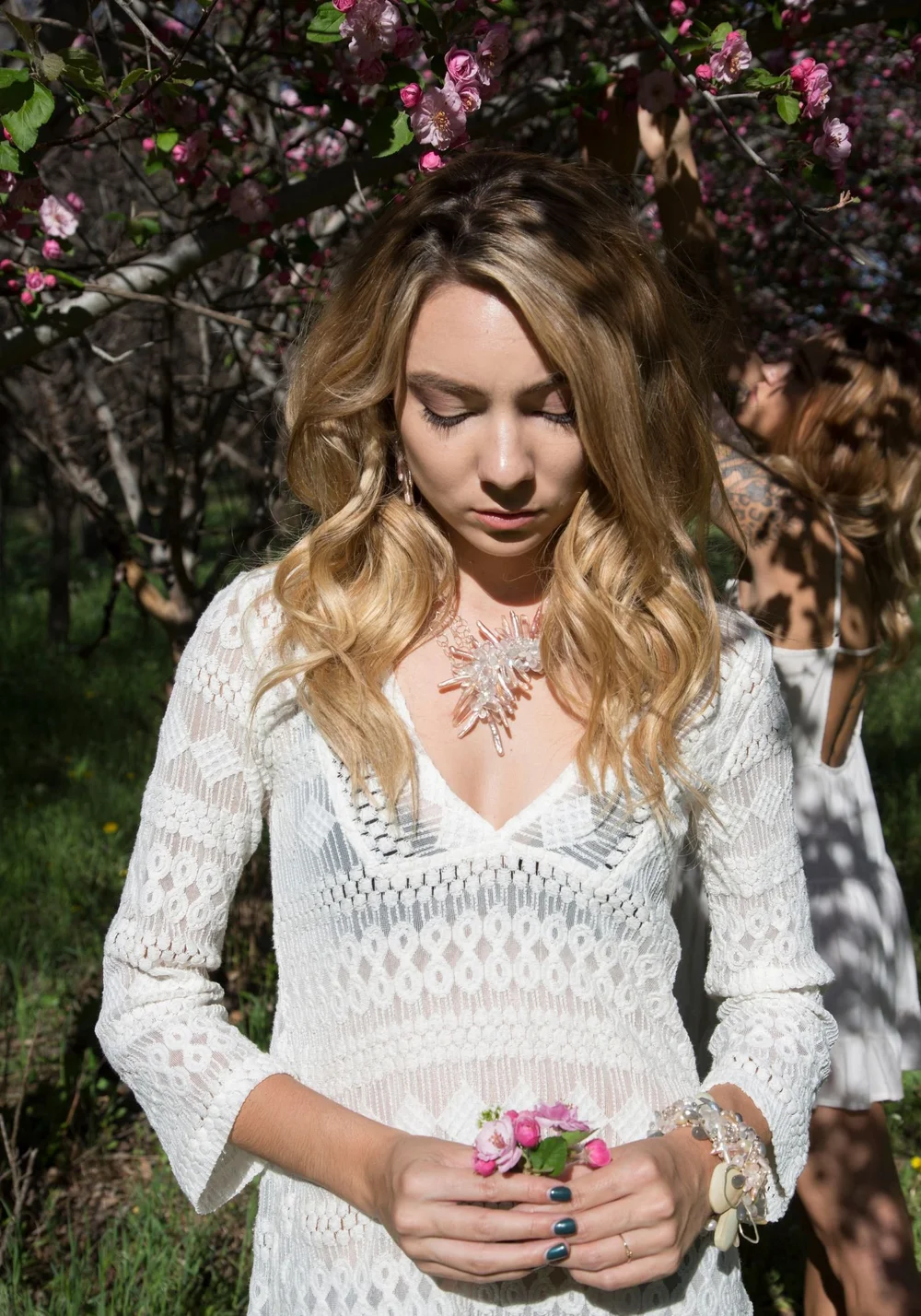 A model in a white lace dress holding pink blossoms, wearing a crystal starburst necklace and a gemstone bracelet