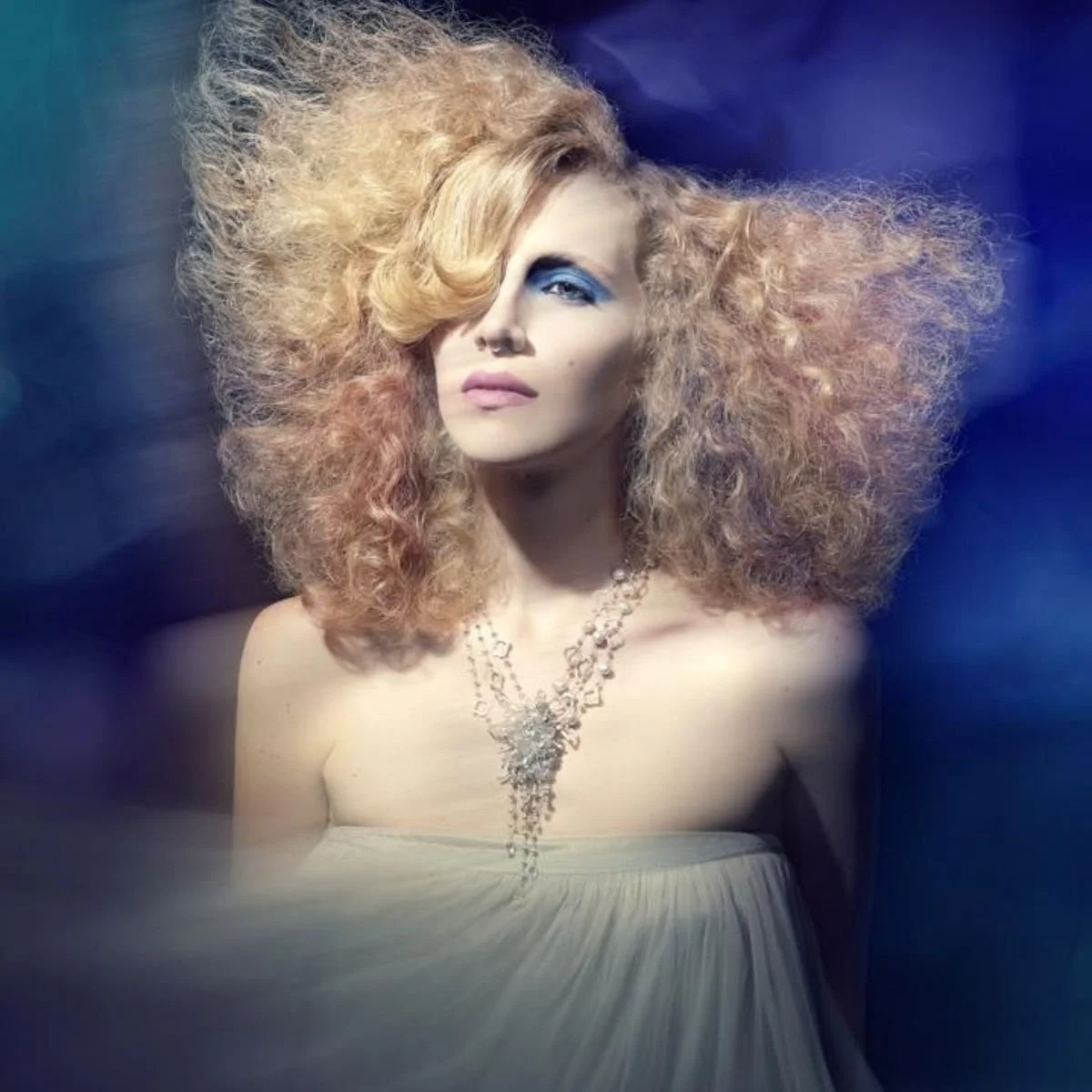 A model with voluminous blonde hair against a blue backdrop wearing a crystal statement necklace