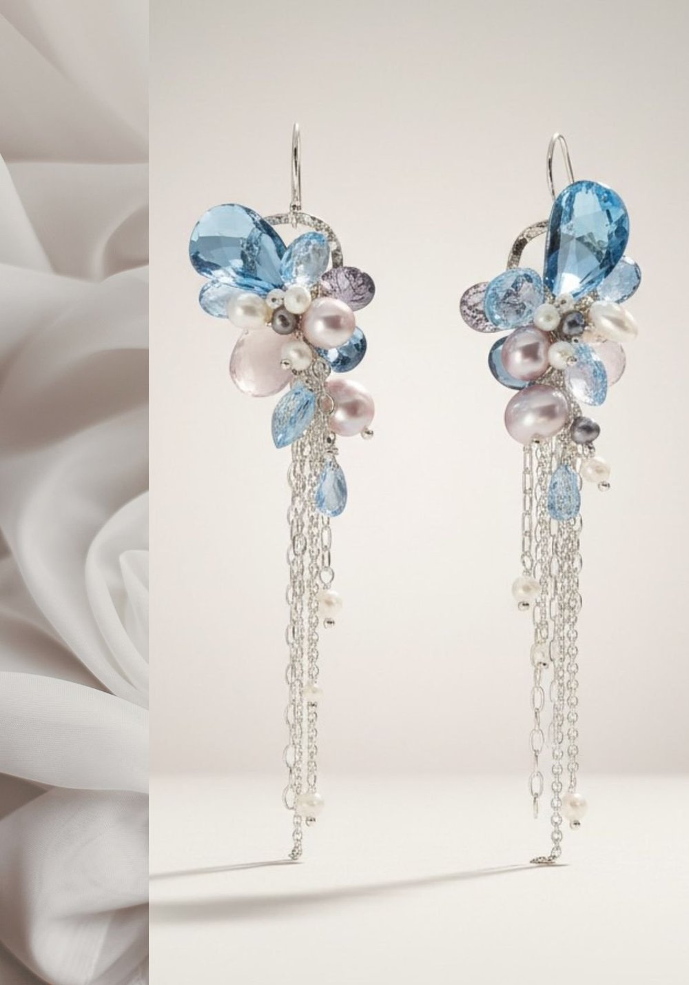 Silver earrings with clusters of pale blue faceted stones and lavender pearls above long silver chain fringe, on a white backdrop
