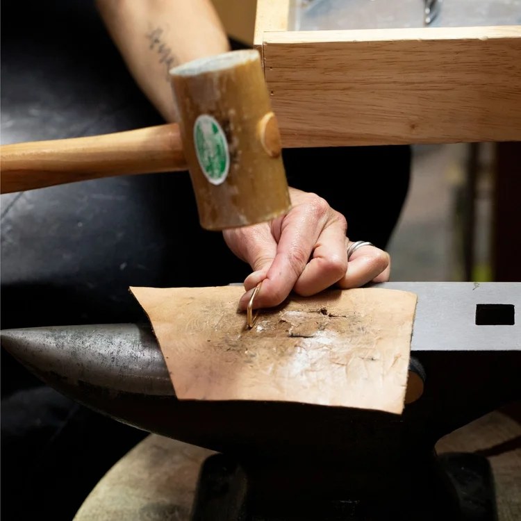 Andrea Li's hands holding a rawhide mallet over a steel block, shaping metal on leather
