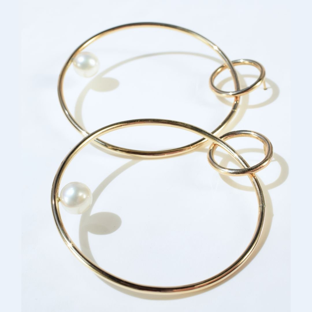 A pair of large gold hoop drop earrings with pearl captured in natural light, emphasizing their shimmer and unique sculptural form