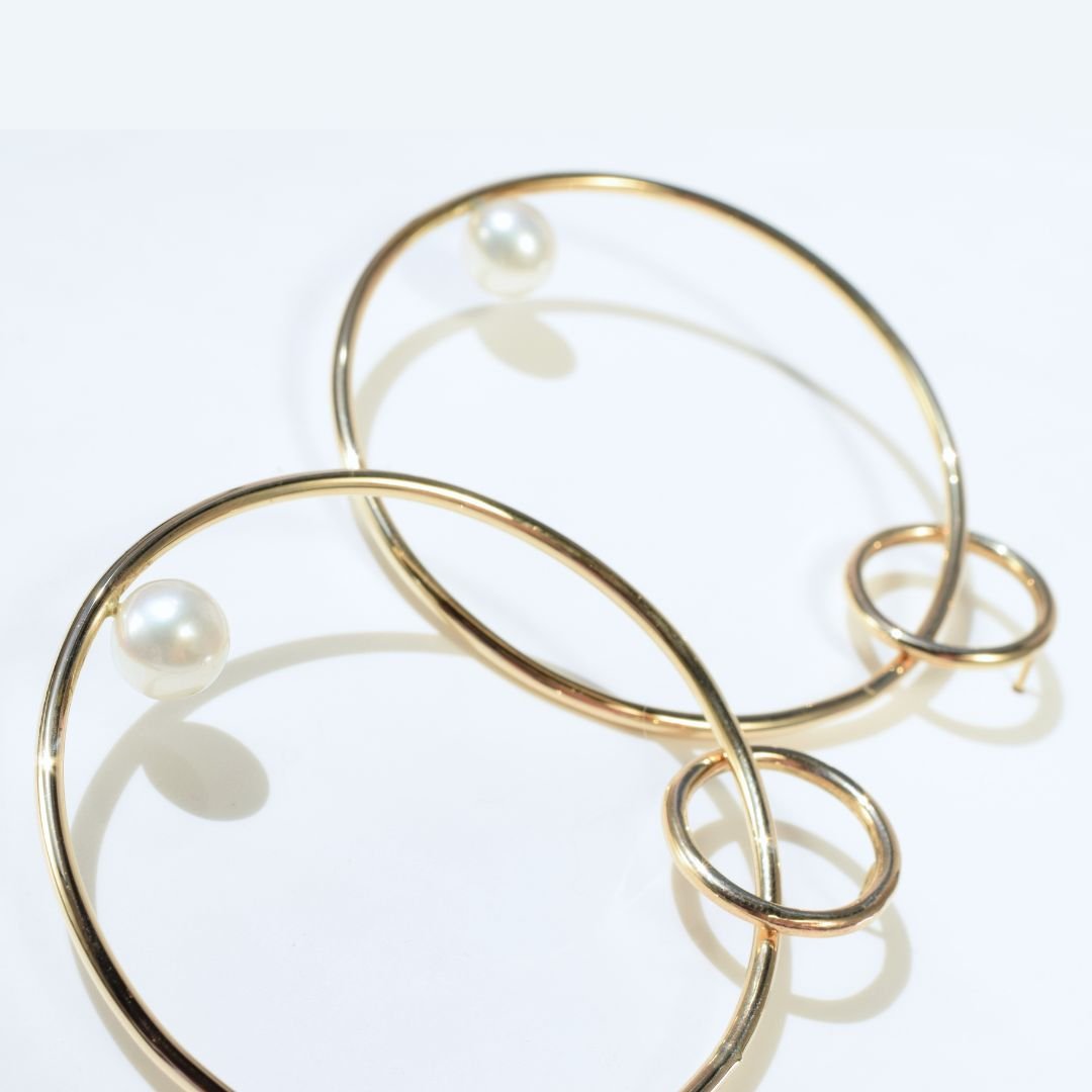 A pair of large gold hoop drop earrings with pearl captured in natural light, emphasizing their shimmer and unique sculptural form