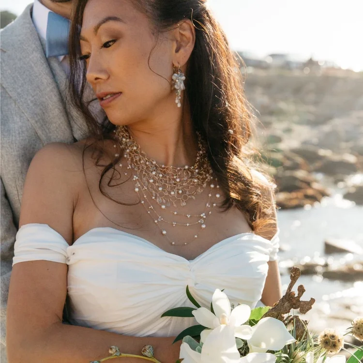 A bride in an off-shoulder gown and a fine beaded necklace beside the groom by the sea