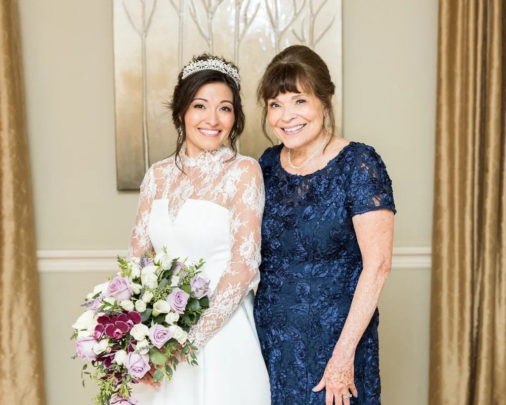 A bride in a jeweled tiara and high-neck lace gown beside her mother in a navy lace dress, a pink and purple bouquet in hand