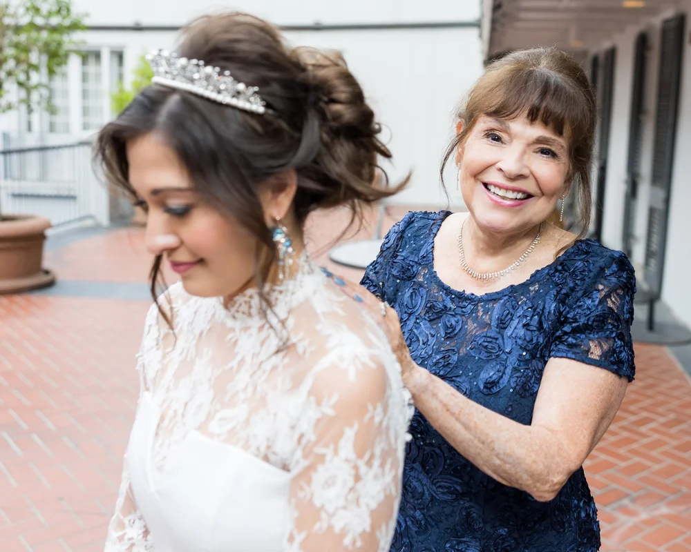 A smiling mother in a navy lace dress adjusting the lace at the bride's shoulder, the bride in a tiara and drop earrings