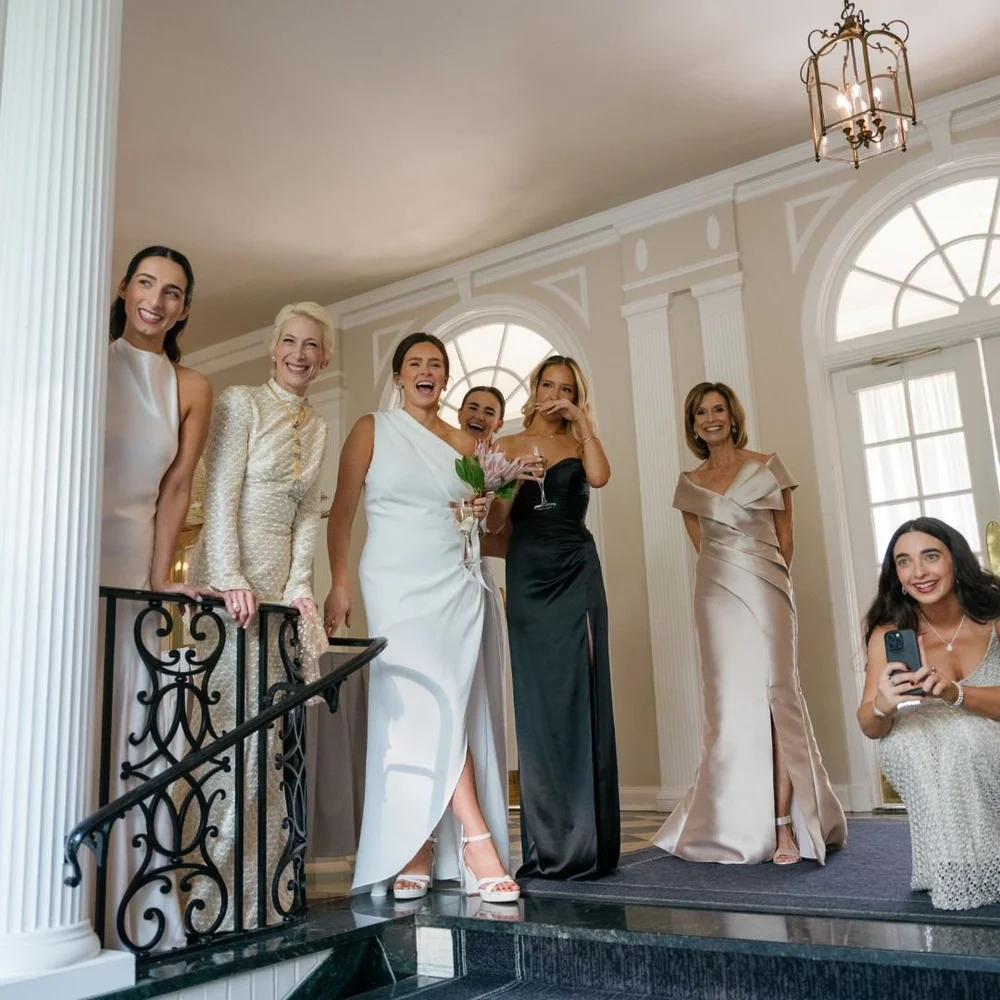 Women in long gowns gathered at the top of a staircase, one taking a phone photo