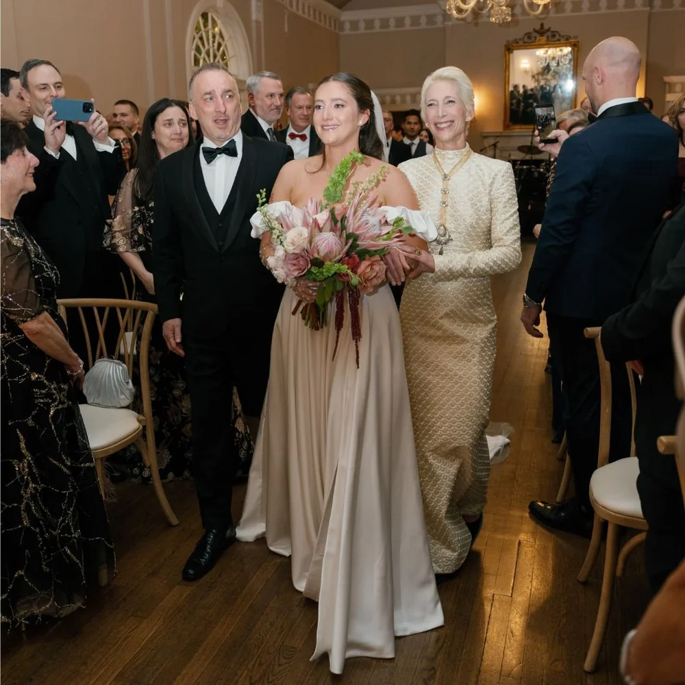 A bride in a blush gown walking the aisle between her father in a tuxedo and her mother in an ivory and gold coat