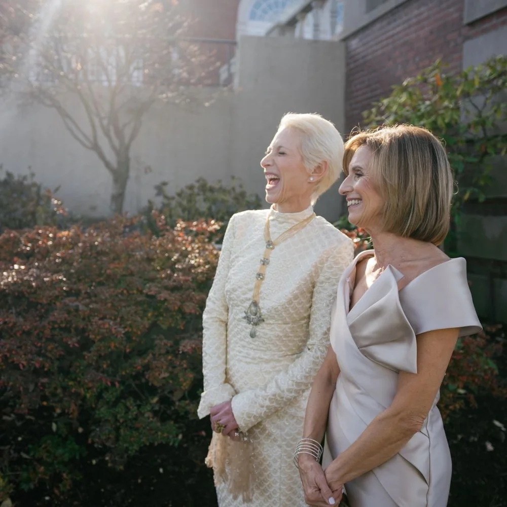 A woman with white hair in an ivory and gold jacket and pendant laughing beside a woman in a champagne off-shoulder dress