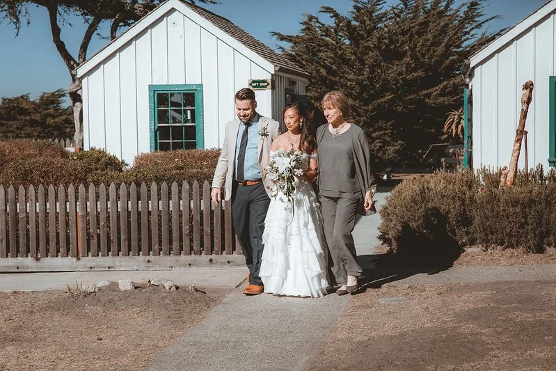 A bride and groom walking out of a white cottage with a woman in a gray outfit beside them