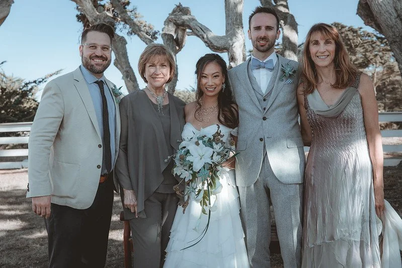 A wedding party of five posed outdoors under cypress trees, the bride holding white lilies