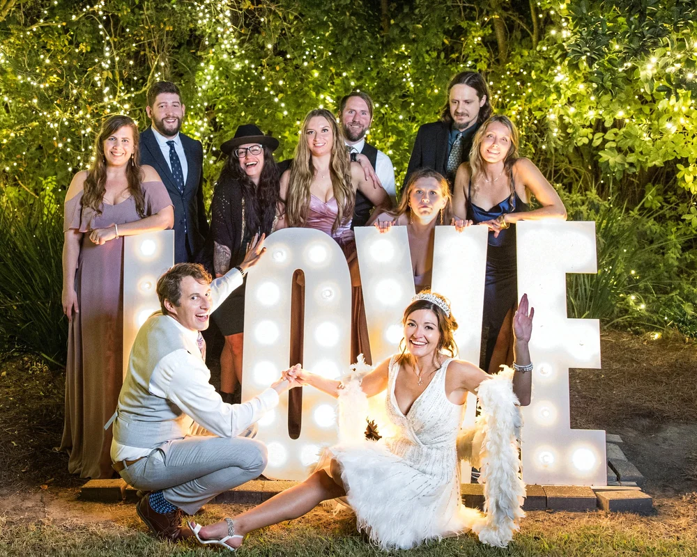Wedding guests posed behind glowing LOVE marquee letters, the couple seated in front