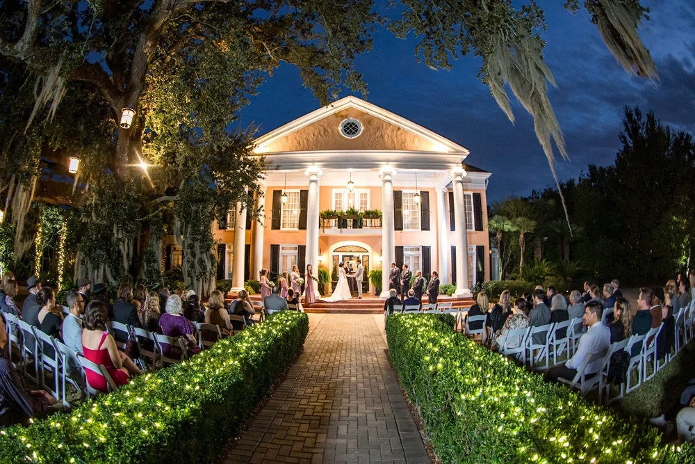 A columned house lit at night above a hedge-lined aisle of seated wedding guests