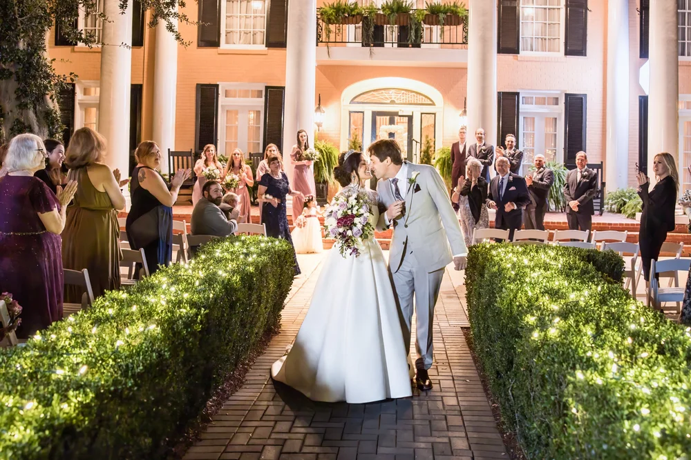 A bride and groom kissing in a hedge-lined aisle as guests applaud before a columned house
