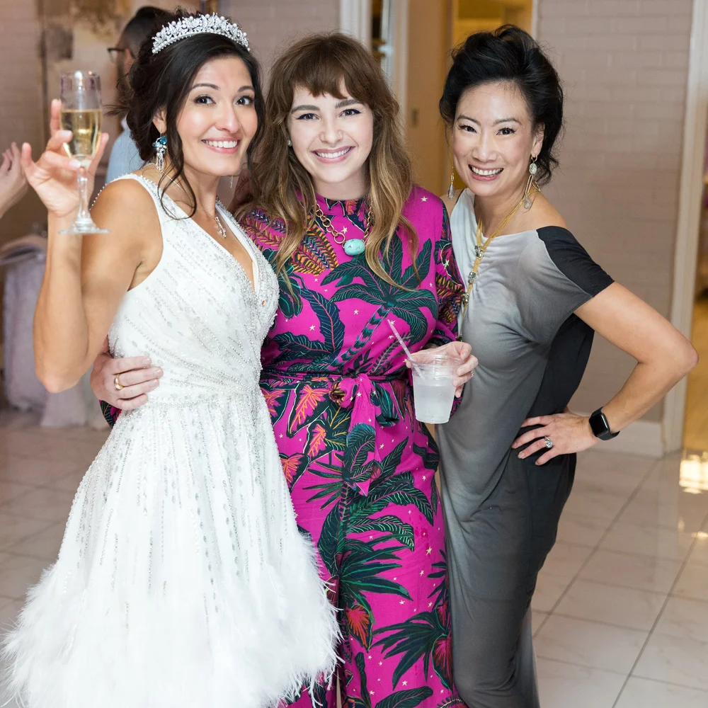 A bride in a tiara with two guests, one in a pink and green print dress, smiling with drinks