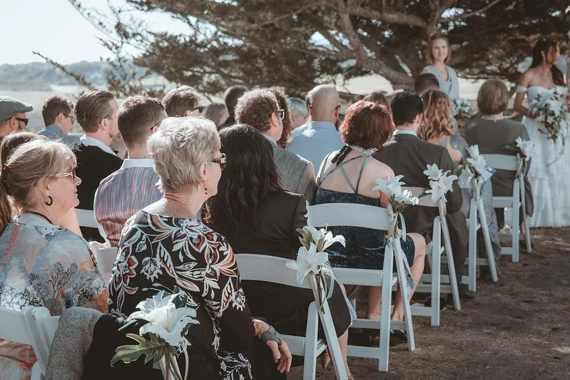 Guests seated in white chairs at an outdoor ceremony under cypress trees