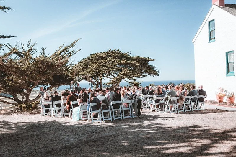 Guests seated for a seaside ceremony beside a white cottage under cypress trees
