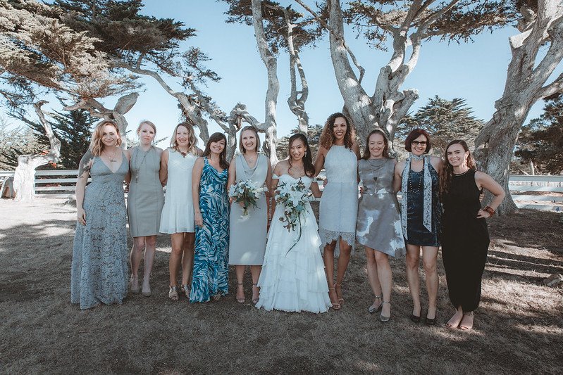 Nine women in blue, gray and white dresses posed with the bride under cypress trees