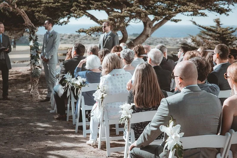 Guests watching an outdoor ceremony beneath cypress trees by the sea