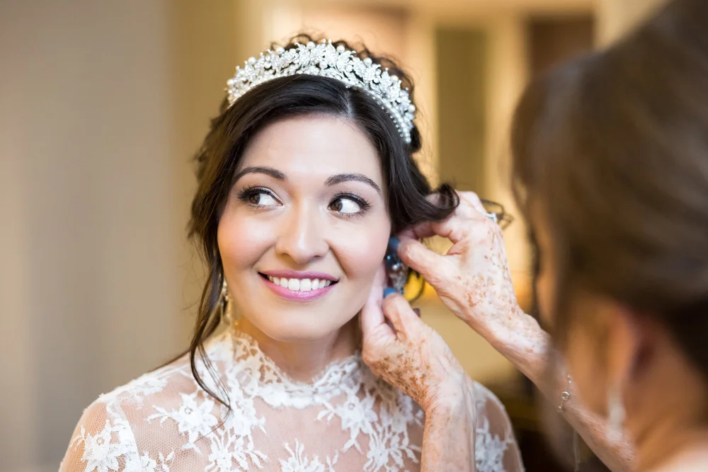 A bride in a jeweled tiara and lace gown smiling as blue drop earrings are fastened