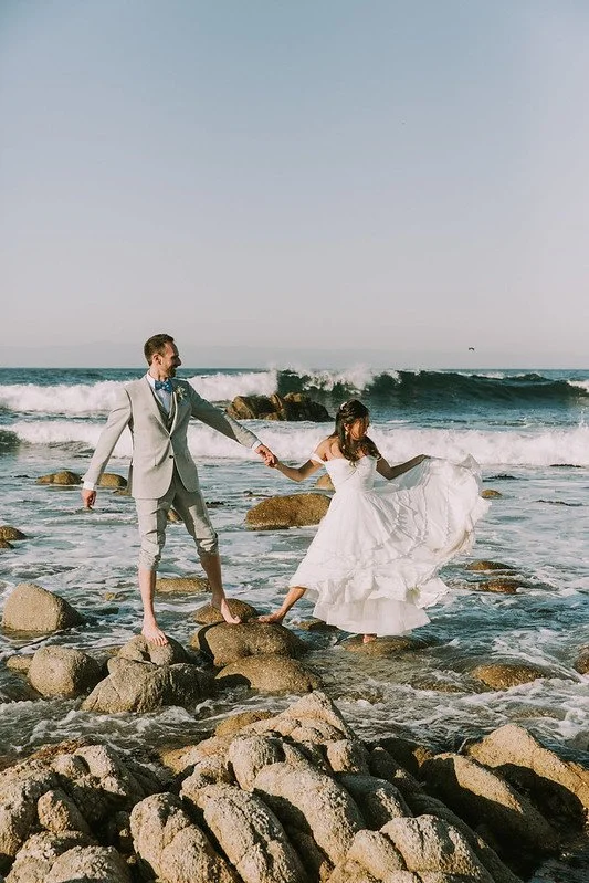 A bride holding her skirt and the groom's hand as they cross wet rocks at the ocean's edge