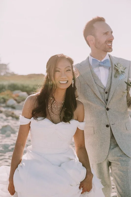 A smiling bride in an off-shoulder gown and a fine beaded necklace beside the groom in a gray suit and blue bow tie