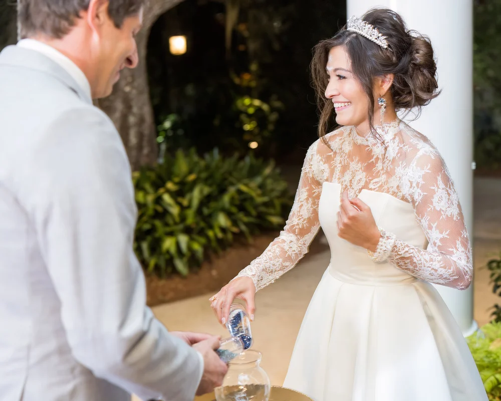 A bride in a tiara, lace gown and blue drop earrings smiling as the groom pours blue sand into a glass vase at night