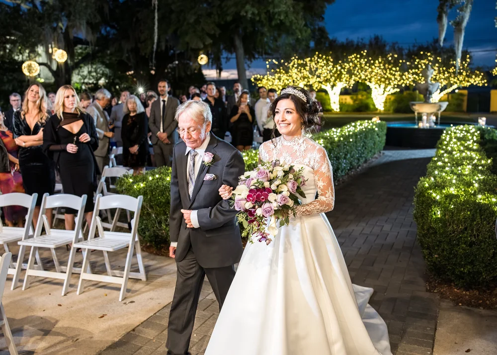 A bride in a jeweled tiara and long-sleeved lace gown walking a lit garden aisle at night on an older man's arm
