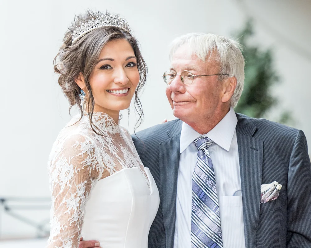 A smiling bride in a tiara and lace gown beside an older man in a gray suit
