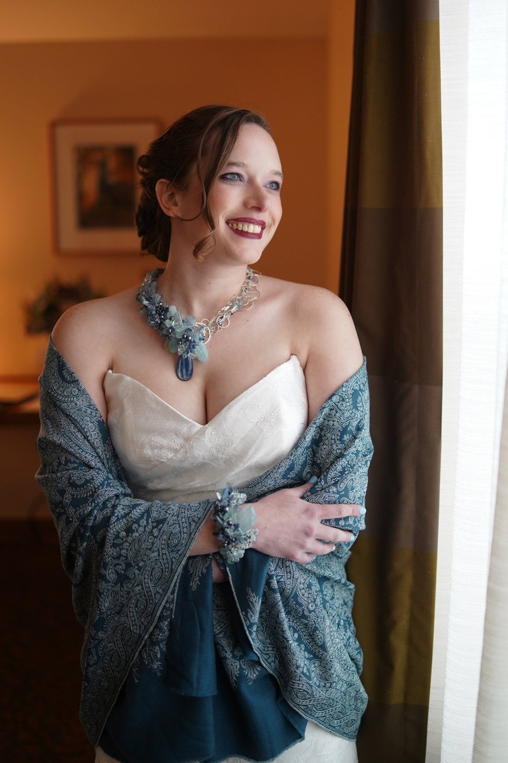 A smiling bride in a strapless gown and blue paisley shawl wearing a blue statement necklace and cuff by a window