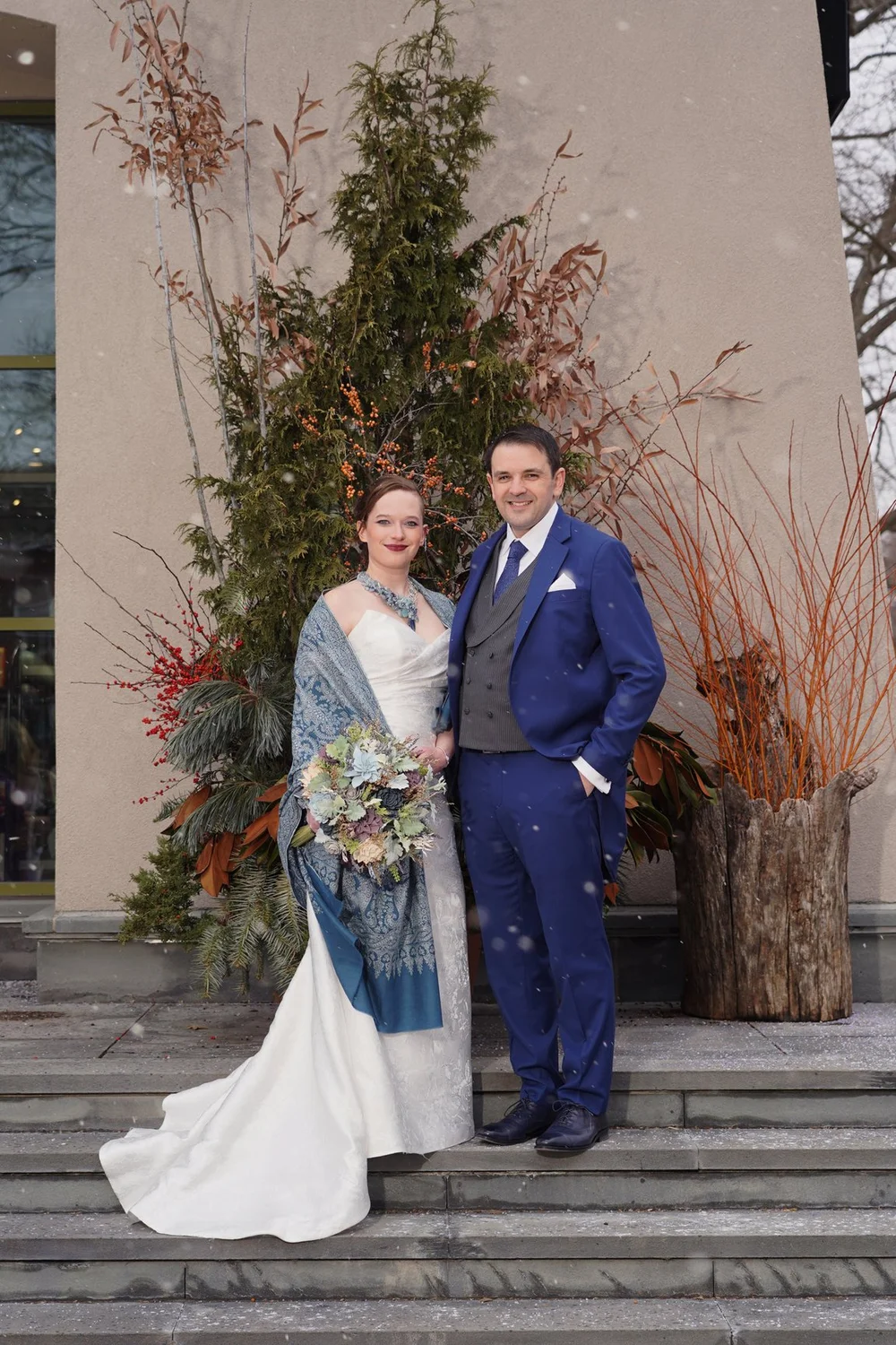 A bride in a blue paisley shawl and the groom in a blue suit on stone steps before a tall winter arrangement