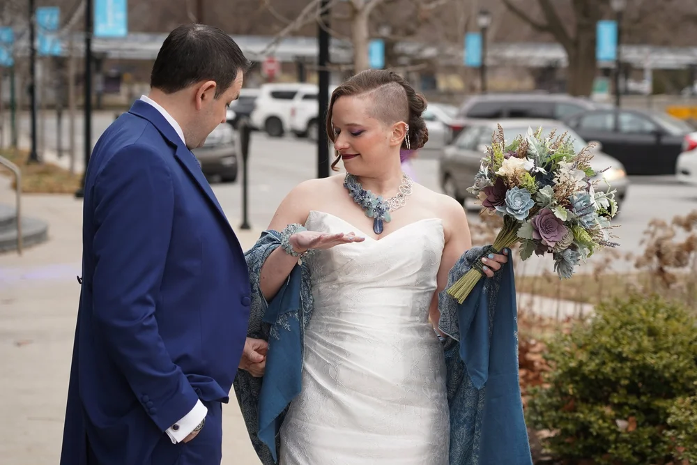 A bride in a strapless gown and blue shawl showing her cuff to the groom in a blue suit, a blue and lavender bouquet in hand