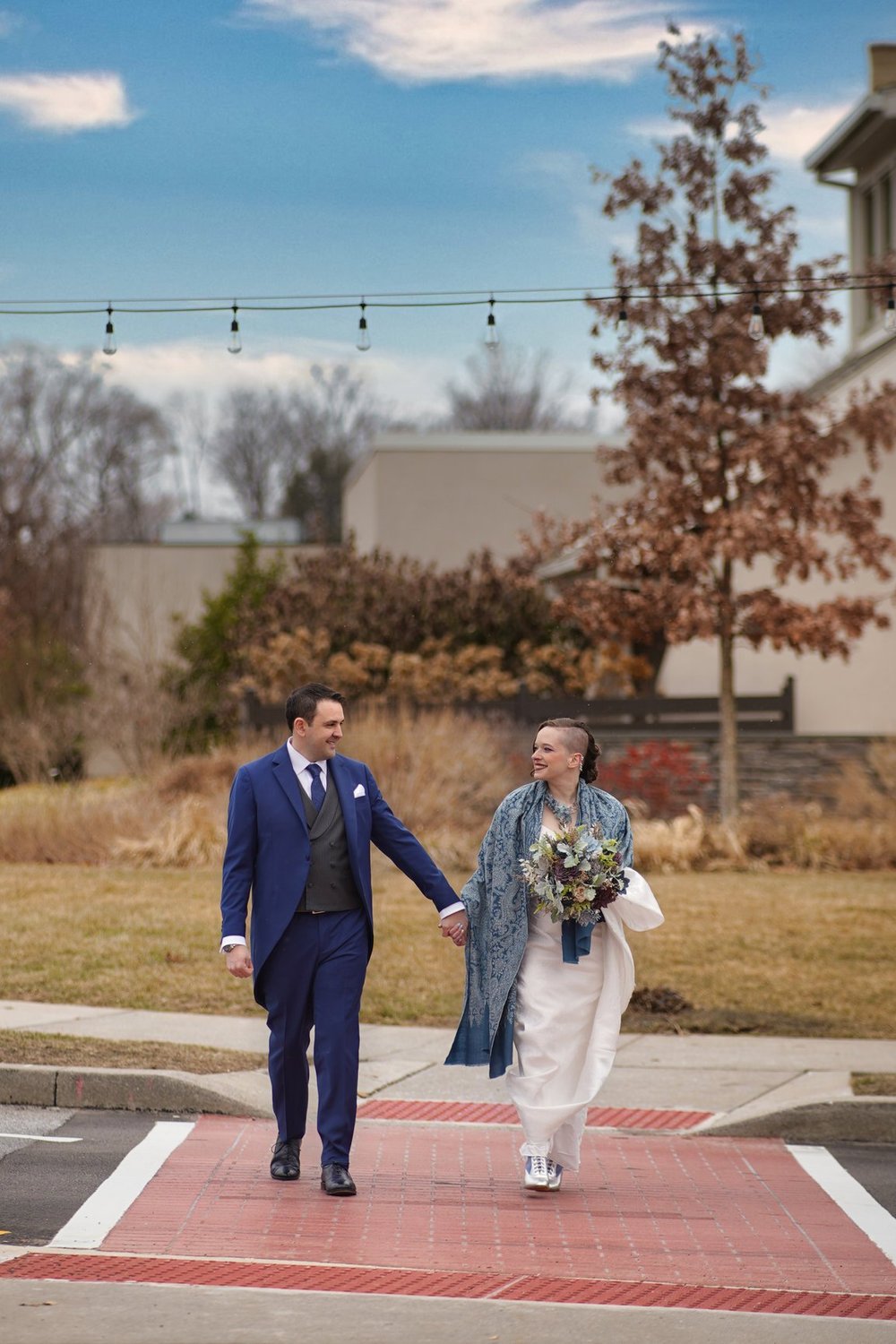 A bride and the groom in a blue suit walking hand in hand across a crosswalk