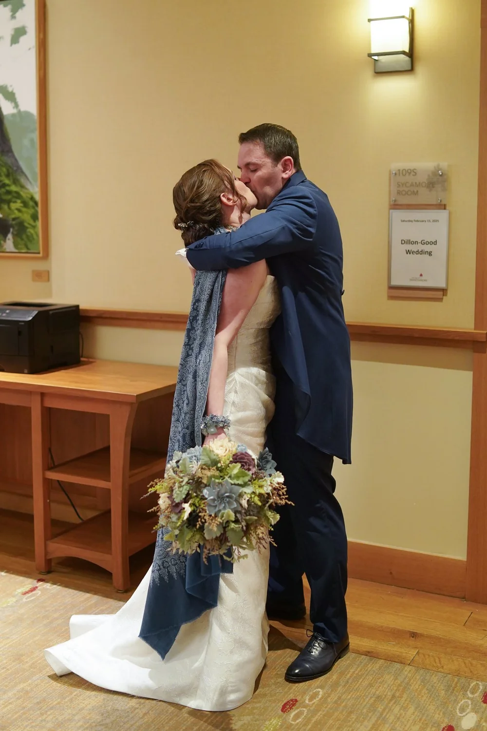 A bride in a blue shawl and the groom in a blue suit kissing indoors, her bouquet in hand