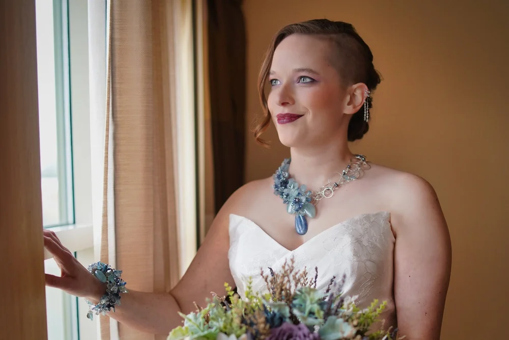 A bride wearing a blue statement necklace and cuff looking out a window, a blue and lavender bouquet in hand