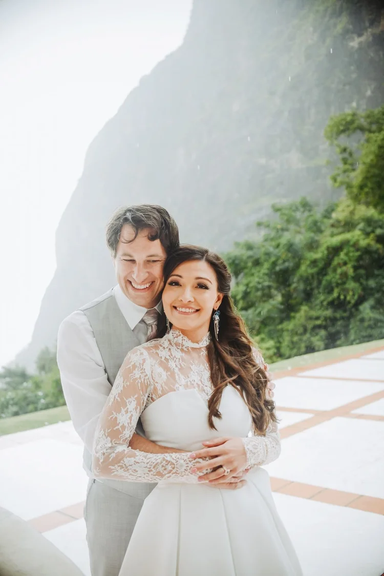 A bride in a high-neck lace gown and the groom in a gray vest embracing on a terrace below a green mountainside
