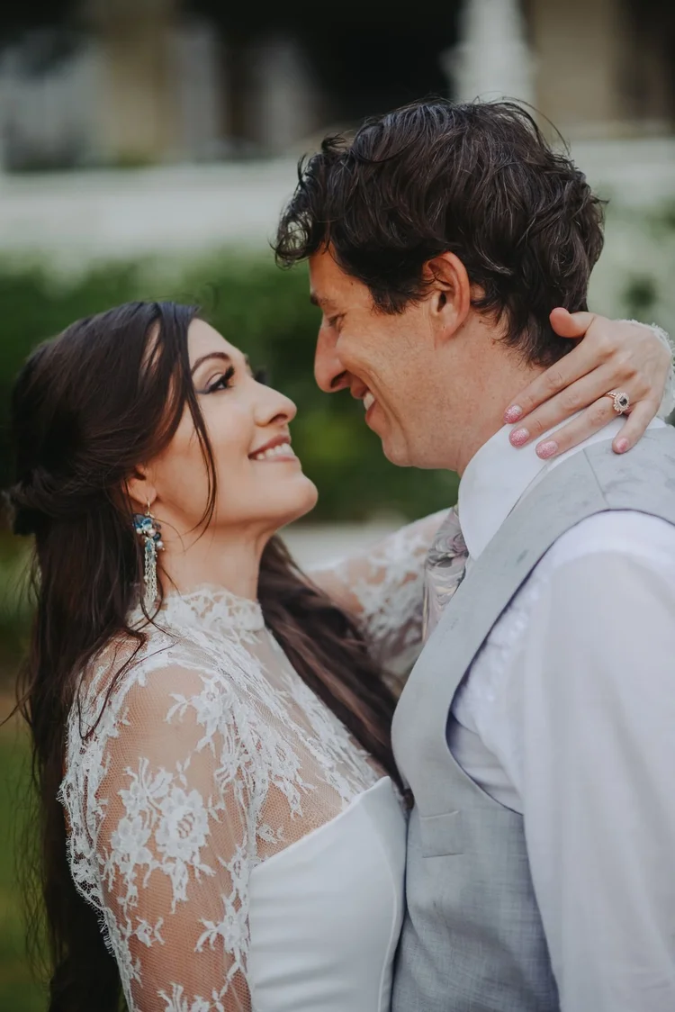 A bride in a high-neck lace gown and drop earrings smiling up at the groom in a gray vest