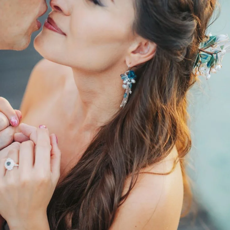 A close view of a bride wearing blue drop earrings and a blue gemstone hair piece, the groom's lips at her cheek