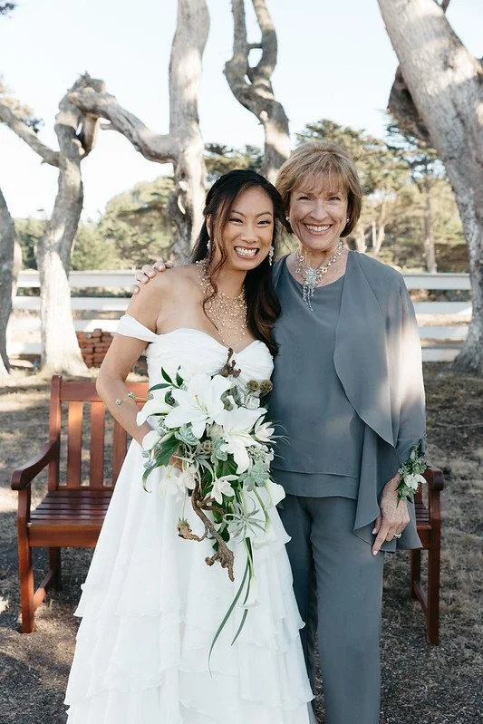 A bride holding white lilies beside an older woman in a gray outfit outdoors
