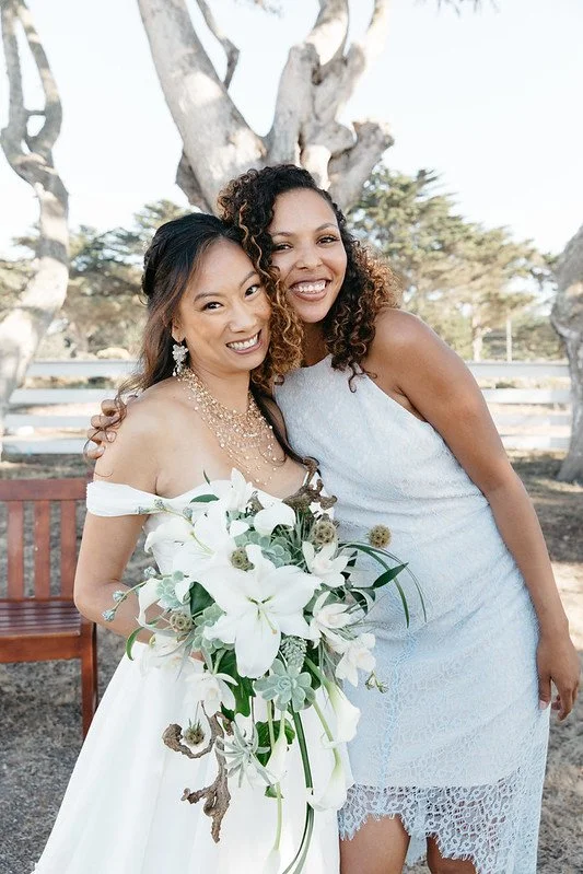 A bride holding white lilies embracing a smiling guest in a white lace dress