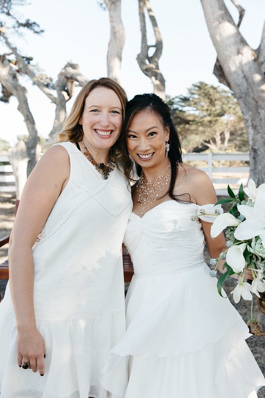A bride and a smiling guest in white posed together under cypress trees