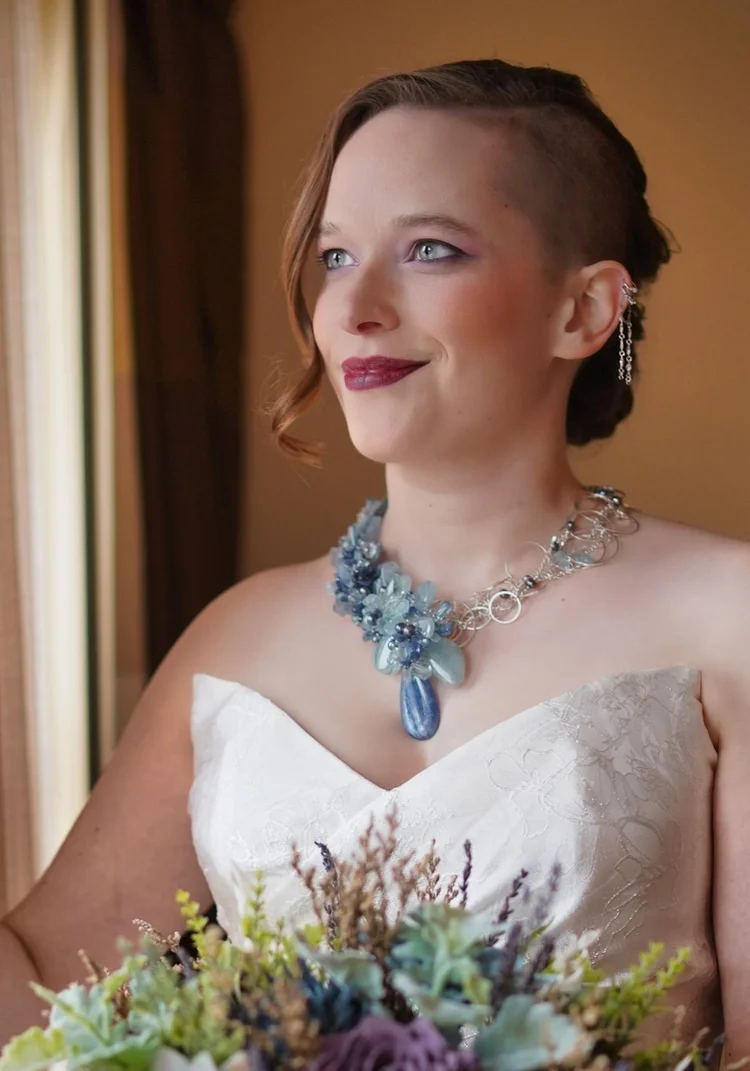 A bride with short red hair wearing a blue statement necklace, a bouquet in hand