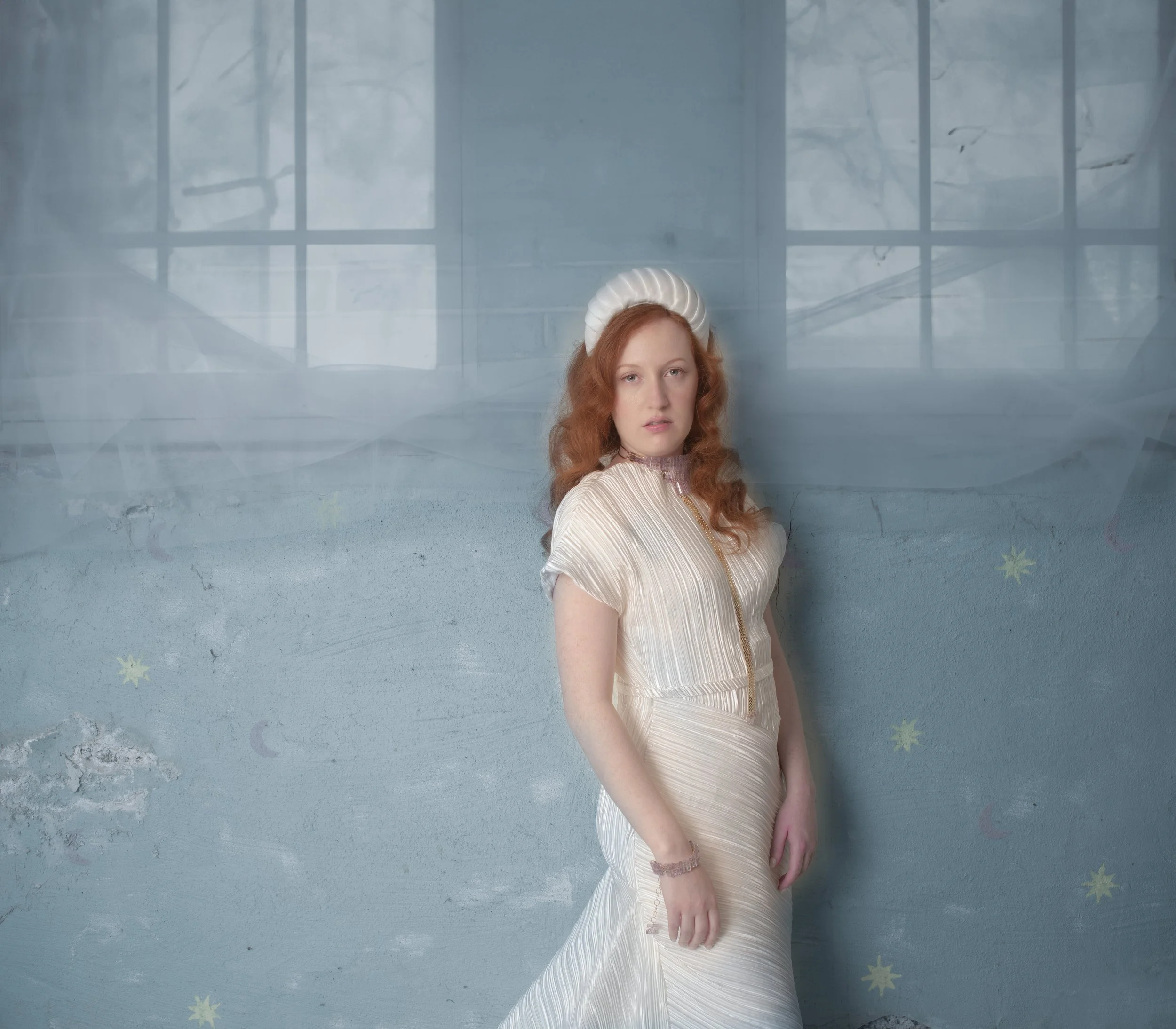 Full-length portrait of a red-haired woman in a flowing white gown, standing barefoot against a dreamy pale blue backdrop with sheer drapery.