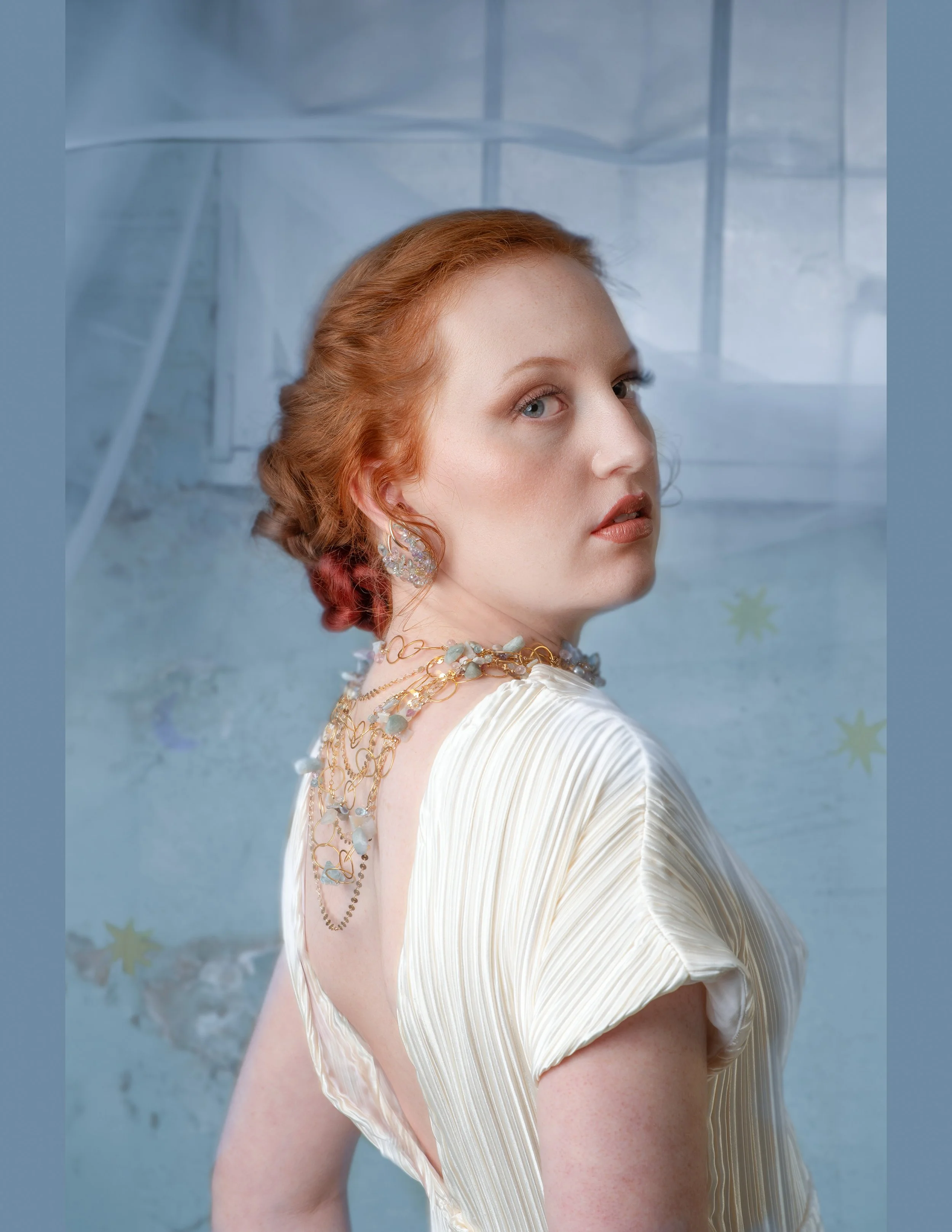 Full-length portrait of a red-haired woman in a flowing white gown, standing barefoot against a dreamy pale blue backdrop with sheer drapery.