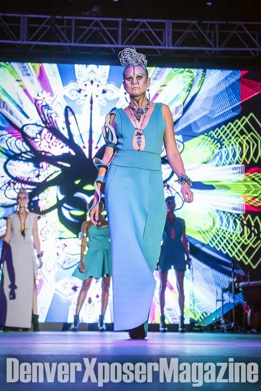 Runway model in a turquoise sculptural gown and futuristic jewelry walks before a vivid neon screen at the Alien Life fashion show.