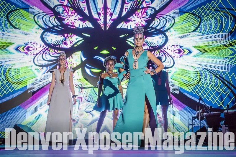 Runway model in a turquoise sculptural gown and futuristic jewelry walks before a vivid neon screen at the Alien Life fashion show.