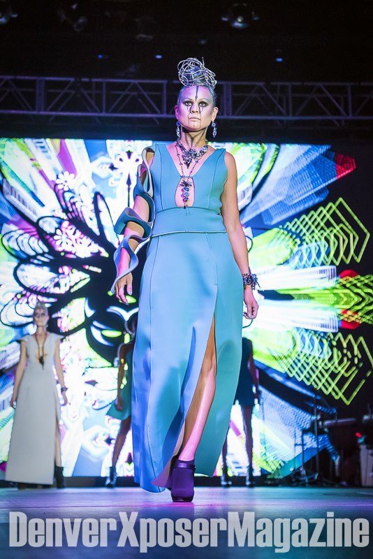 Runway model in a turquoise sculptural gown and futuristic jewelry walks before a vivid neon screen at the Alien Life fashion show.