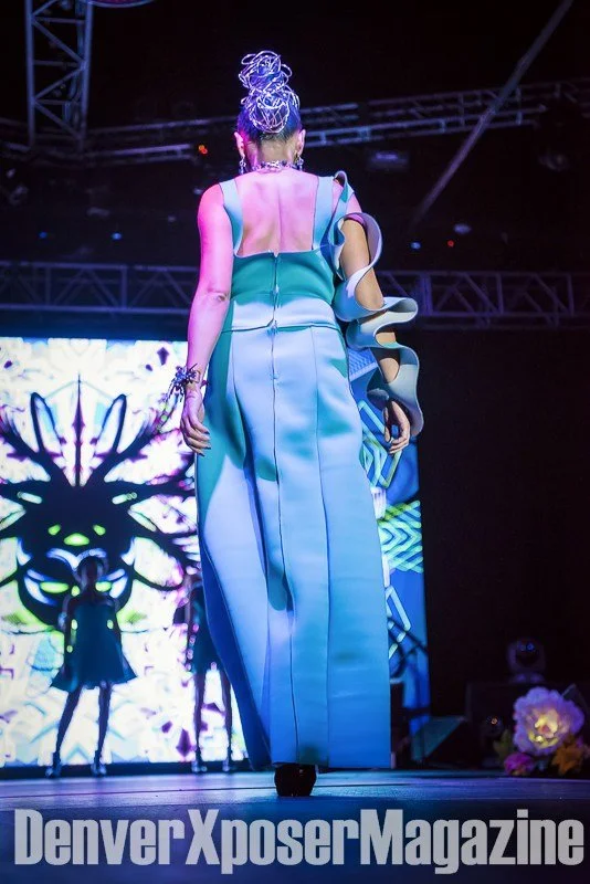 Runway model in a turquoise sculptural gown and futuristic jewelry walks before a vivid neon screen at the Alien Life fashion show.