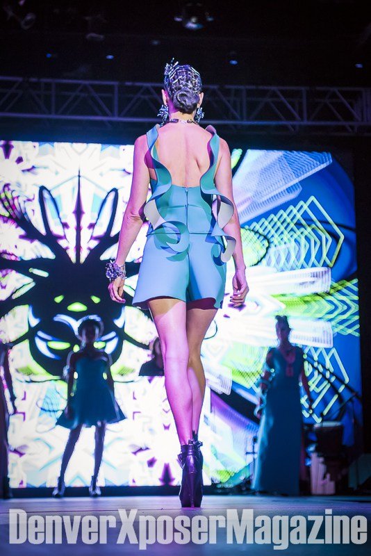 Runway model in a turquoise sculptural gown and futuristic jewelry walks before a vivid neon screen at the Alien Life fashion show.
