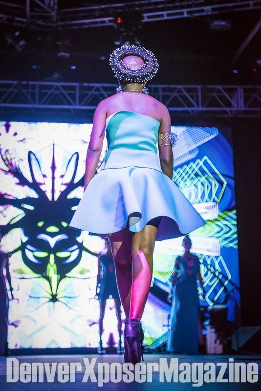 Runway model in a turquoise sculptural gown and futuristic jewelry walks before a vivid neon screen at the Alien Life fashion show.